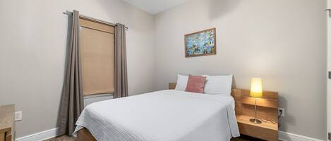 A bedroom with a bed, a nightstand, a lamp, a ceiling fan, a window with curtains, and a painting on the wall.