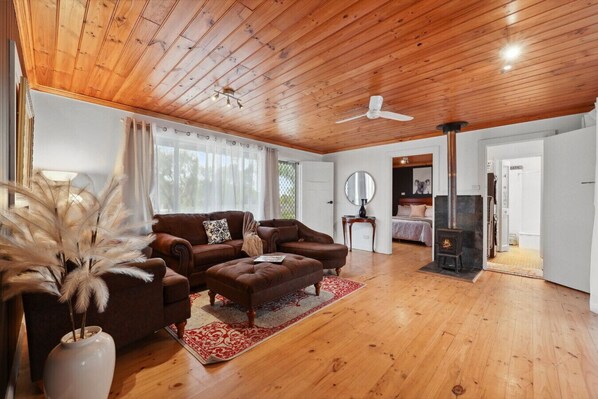 A spacious living room with a wooden ceiling, a sofa set, a fireplace, and a view into the bedroom.