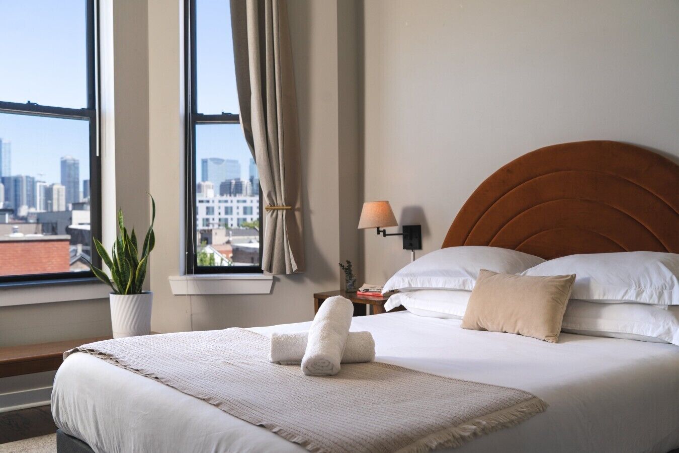 A hotel room with a large bed, a view of the city through a window, a potted plant, and a nightstand with a lamp.