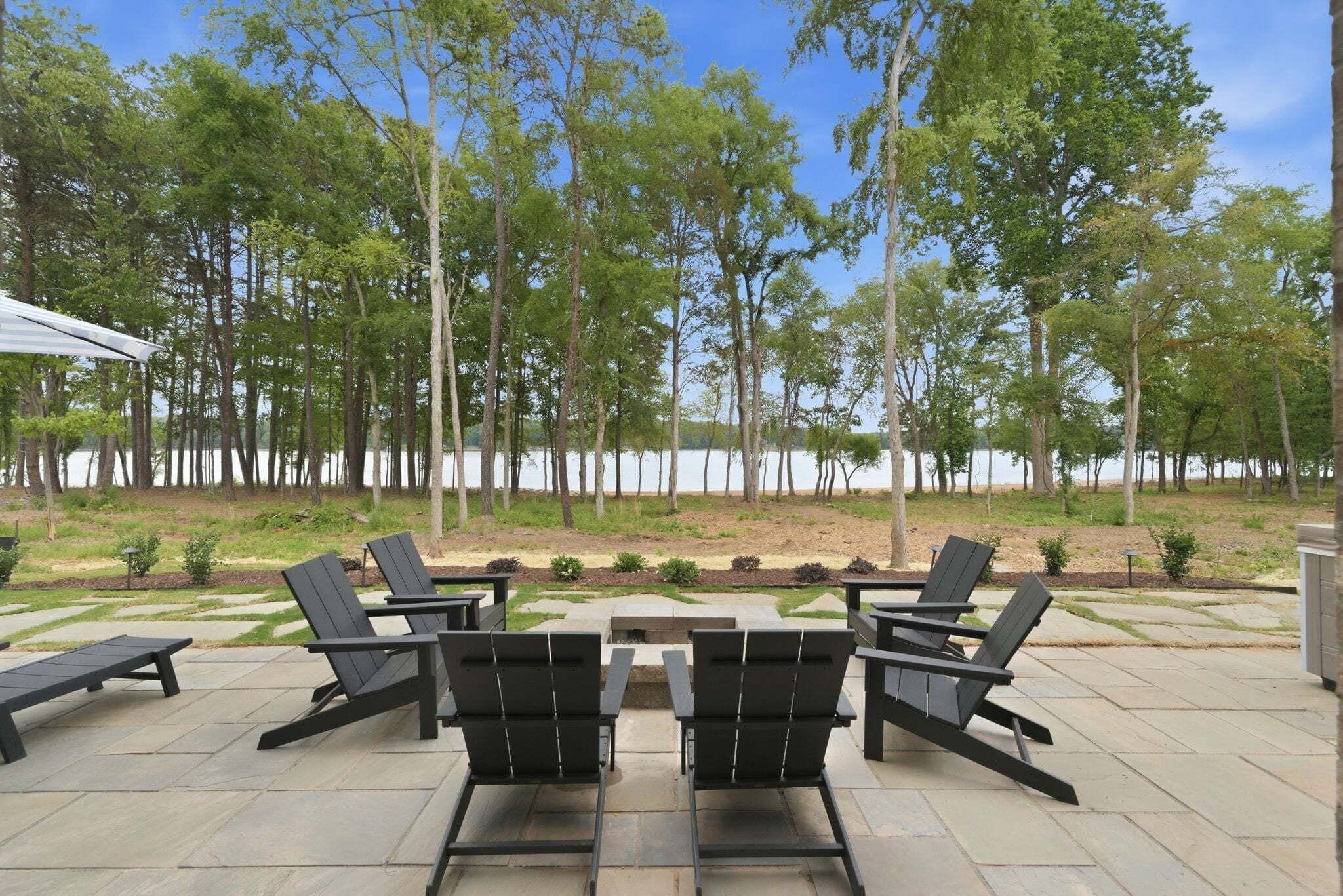 An outdoor patio with four Adirondack chairs, a stone fire pit, and a view of a lake surrounded by trees.