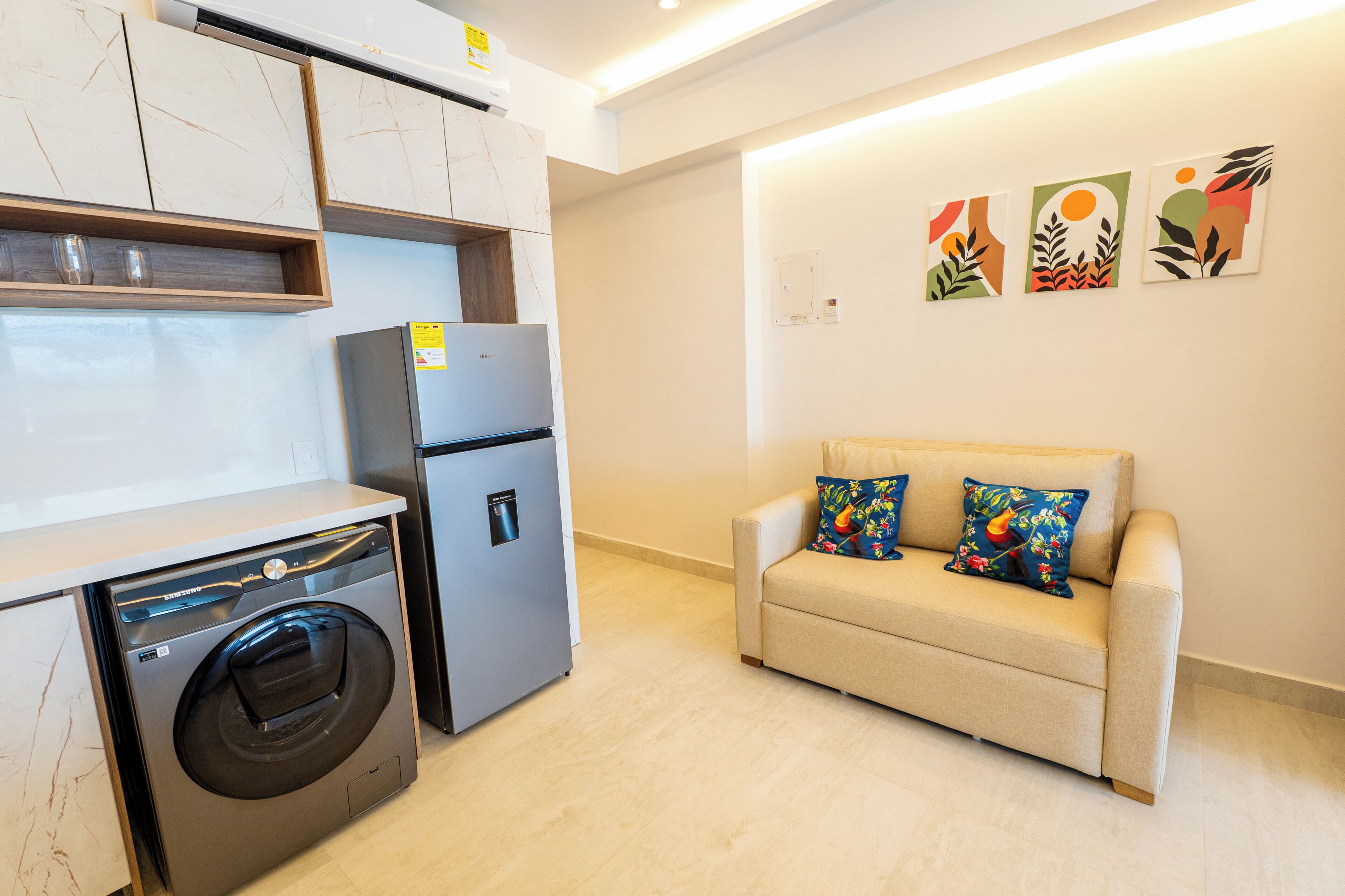 A compact living space with a washing machine, a refrigerator, a sofa with colorful cushions, and abstract wall art.