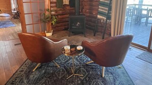 A cozy log cabin interior with two leather chairs, a small round table, a fireplace, and a view of the deck outside. - Edgewater Lodge: Riverfront Family Fun in Grayling (Grayling)
