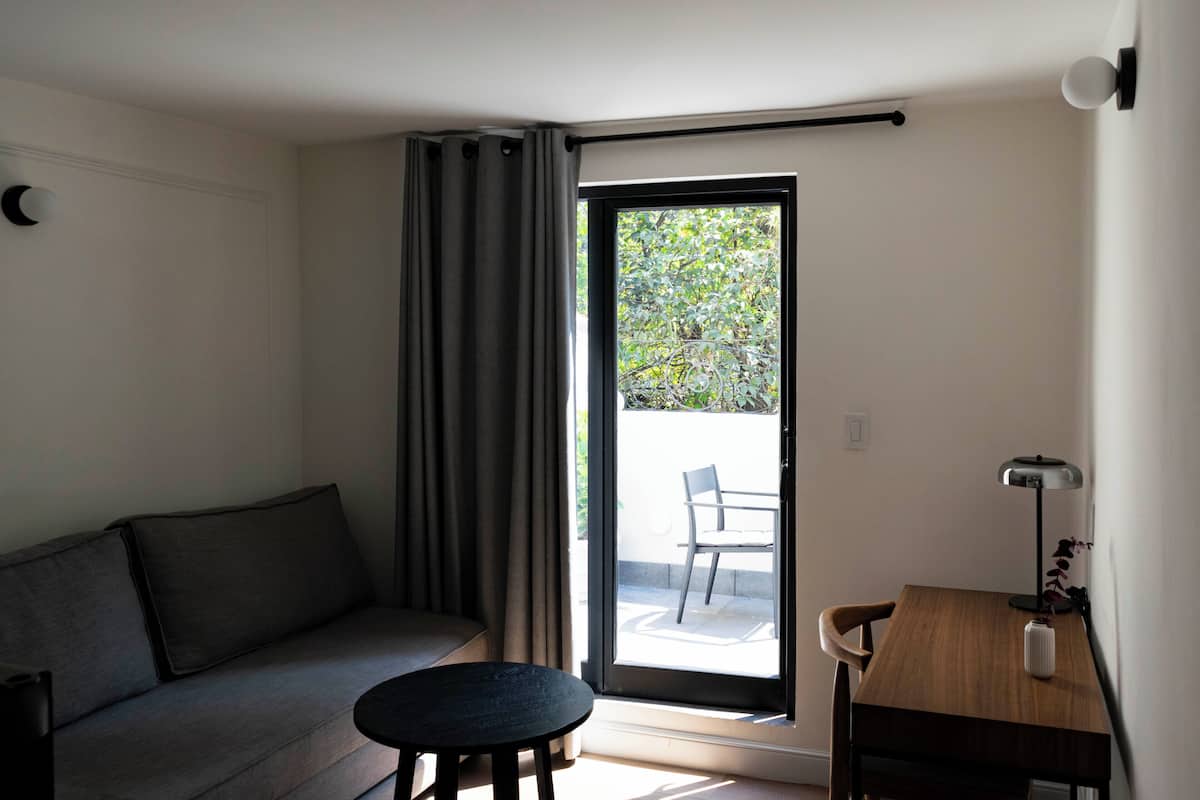 A modern room with a sofa, a small round table, a desk with a lamp, and a sliding glass door leading to an outdoor area.