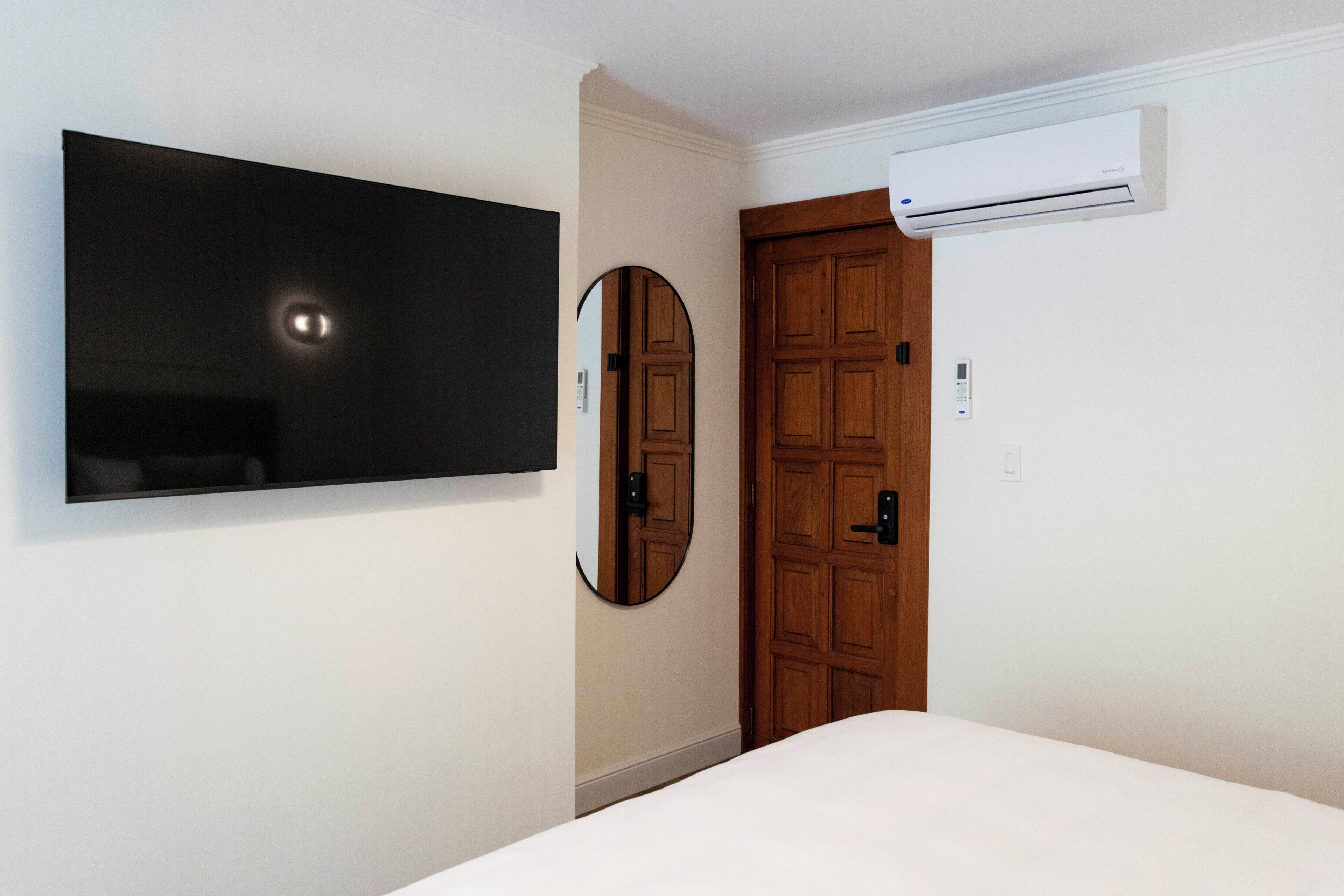 A hotel room with a bed, a mounted TV, a wooden door, and an air conditioning unit.