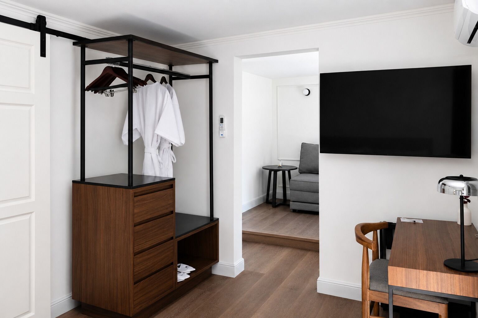 A room with a wooden wardrobe, a desk with a chair, a television, and a grey sofa.