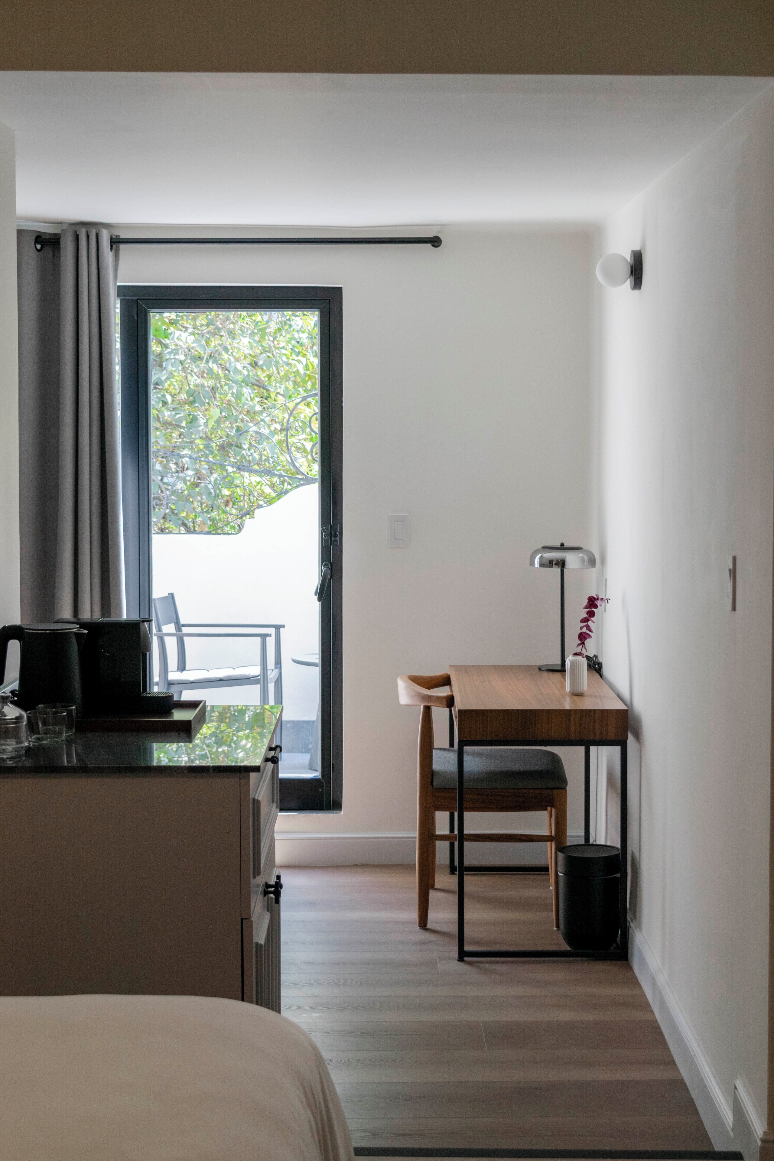 Habitación de hotel con cama, escritorio con silla, mesa con lámpara y vista a un área exterior a través de una puerta.