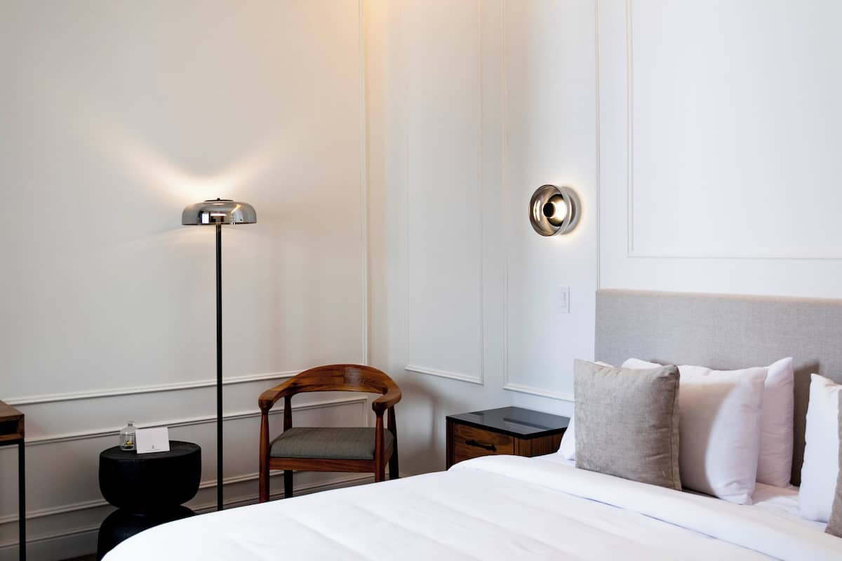 A hotel room with a bed, a chair, a nightstand, a lamp, and a wall-mounted light fixture.