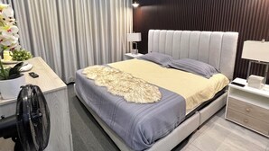 Une chambre d’hôtel moderne, dotée d’un grand lit, d’un bureau avec une chaise, d’un ventilateur sur pied et d’une table de chevet avec une lampe.
