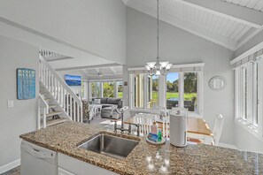 A kitchen with granite countertops, a stainless steel sink, a dining area with a wooden table and chairs, and a staircase with white railings. - Sunsets on Sealoft by Sandpiper | Near Beach (Seabrook Island)
