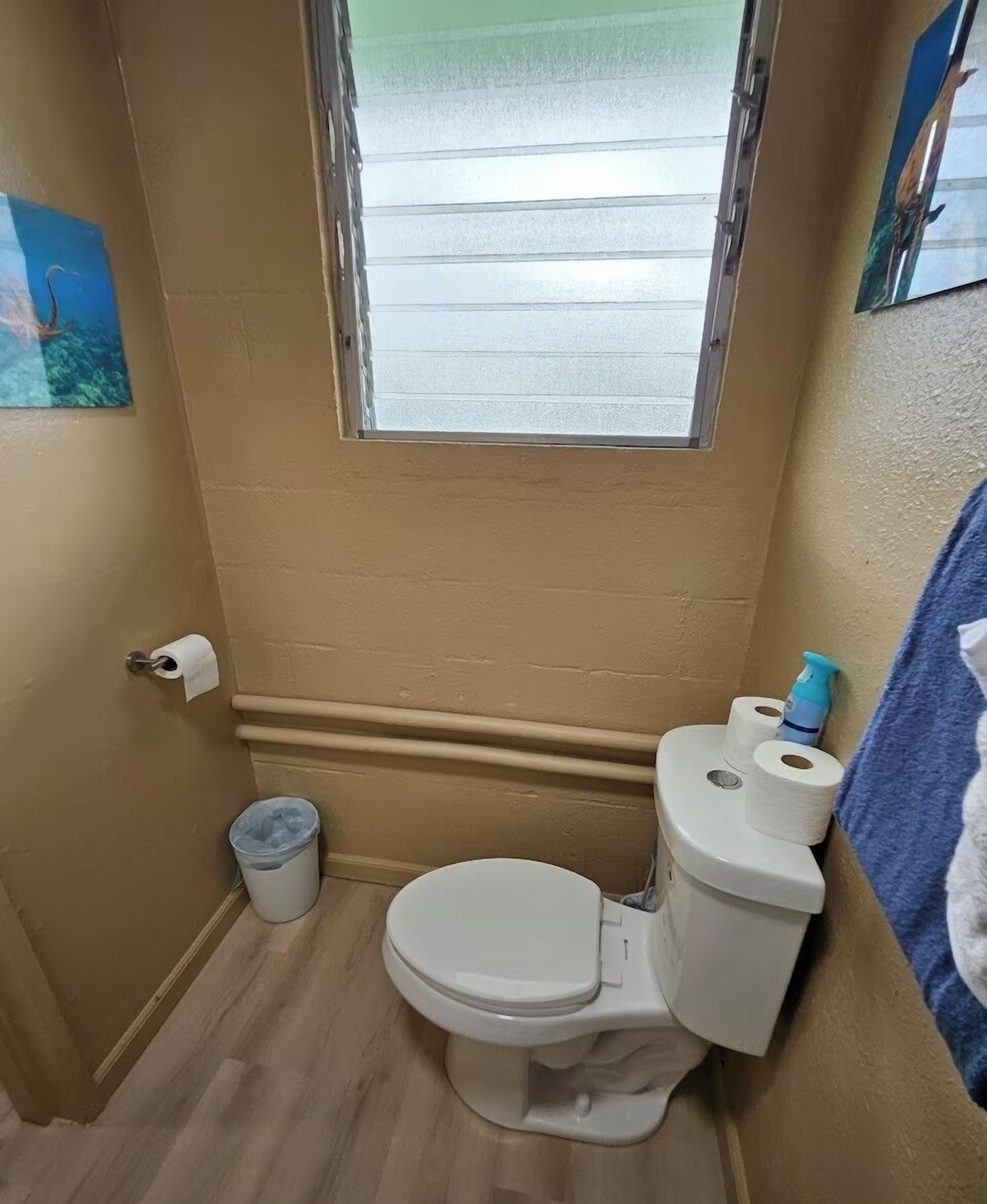 A small bathroom with a toilet, a window, a trash bin, and a towel rack.