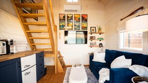Un espace de vie compact comprenant une cuisine, un canapé bleu, un banc blanc, un escalier en bois et un téléviseur fixé au mur.