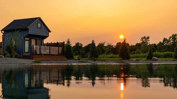 Une maison dotée d’une terrasse offrant une vue sur un lac paisible, entourée d’arbres et illuminée par le coucher du soleil.