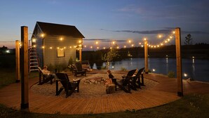 Une terrasse en bois avec des sièges extérieurs et un petit chalet au bord d’un lac, au crépuscule.