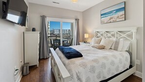A bedroom with a bed, a ceiling fan, a TV, a window with curtains, and a painting on the wall. - Residence 102N at The Sandcastle Condominiums (Wildwood Crest)