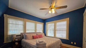 Un dormitorio con cama, mesita de noche, reloj, ventilador de techo y ventanas grandes con persianas.