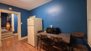 Cocina compacta con un refrigerador blanco, un microondas sobre la encimera y un comedor con una mesa y sillas de madera.