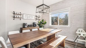 A dining area with a long wooden table, white chairs, a bench, a coffee machine, a plant, and a window with a view of trees. - 5 Bedroom house with amenities galore! Oversized hot tub, outdoor fire pit, smoker and propane grill, sauna, play set, espresso machine, air hockey table, indoor fireplace in both living rooms, and legacy bar. (Colorado Springs)