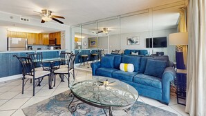 Una sala de estar moderna con una mesa de centro de vidrio, un sofá azul y una cocina visible a través de un separador de vidrio.