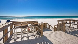 Una terraza de madera con sillas de metal y barandilla de madera que da a una playa.