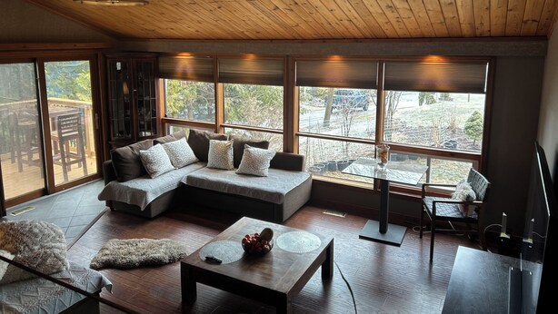 A spacious room with a large sectional sofa, a wooden ceiling, and large windows offering a view of the outdoors.