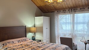 A bedroom with a large bed, a wooden ceiling, a chandelier, a white wardrobe, a desk with a laptop, and a window with sheer curtains.