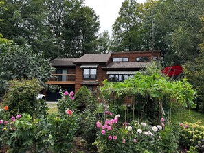 A two-story wooden house surrounded by lush greenery and flowering plants.