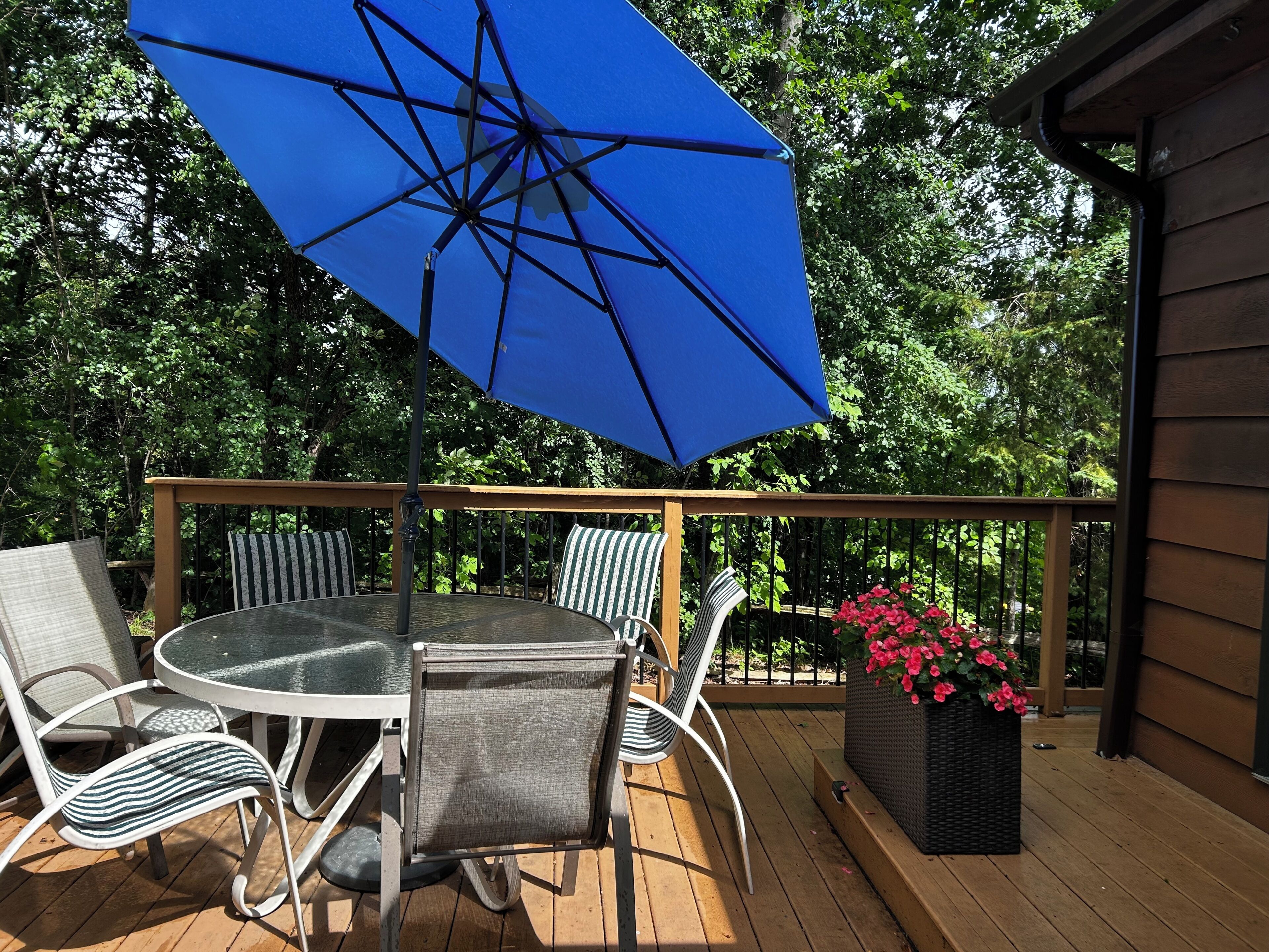 A wooden deck with a round table and chairs, a blue umbrella, and a planter with pink flowers.
