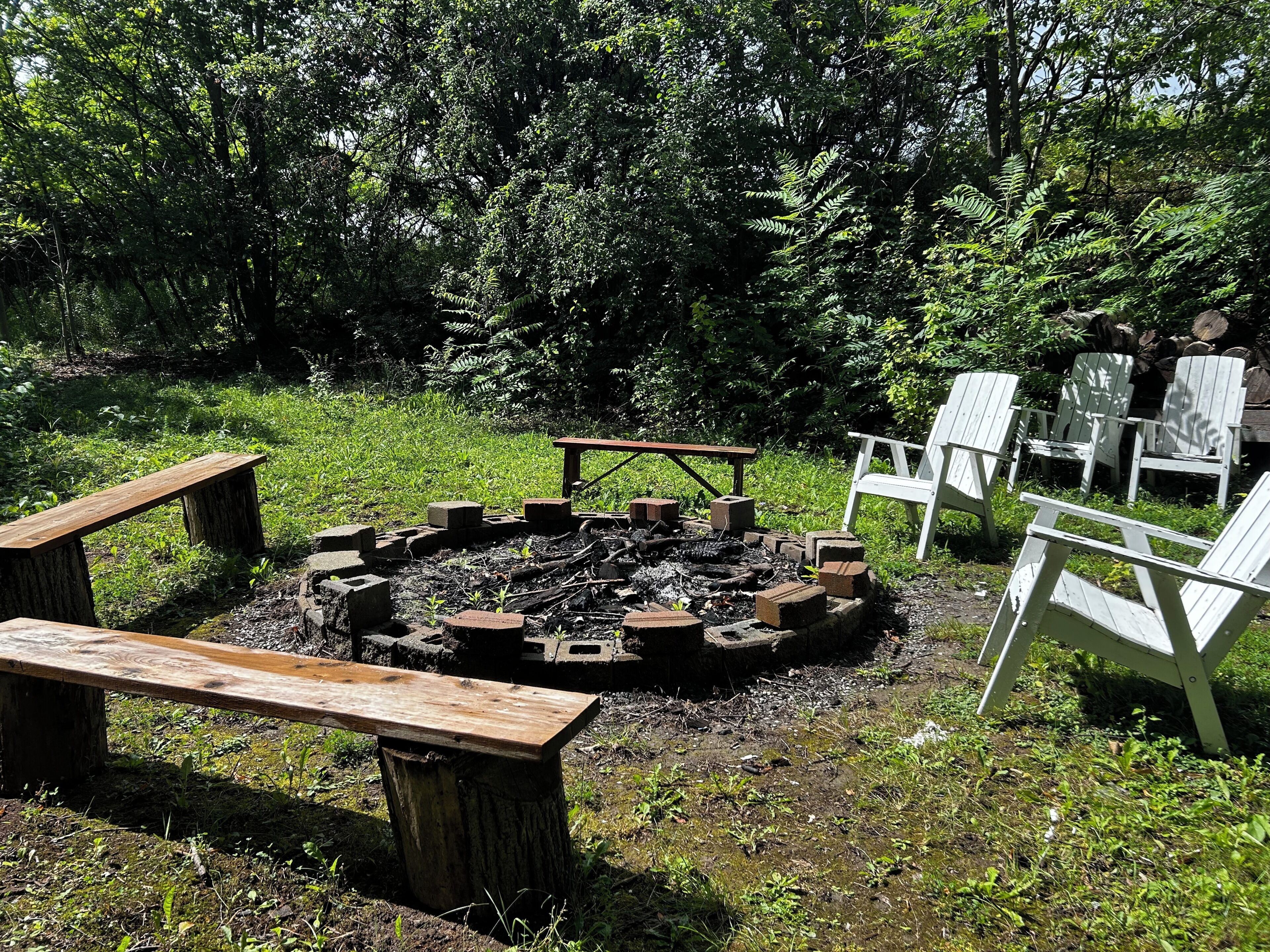 Un foyer circulaire entouré de sièges, notamment de bancs et de fauteuils Adirondack, installé dans un espace verdoyant avec des arbres en arrière-plan.
