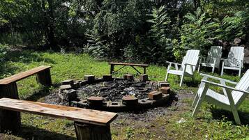 Un foyer circulaire entouré de sièges, notamment de bancs et de fauteuils Adirondack, installé dans un espace verdoyant avec des arbres en arrière-plan.