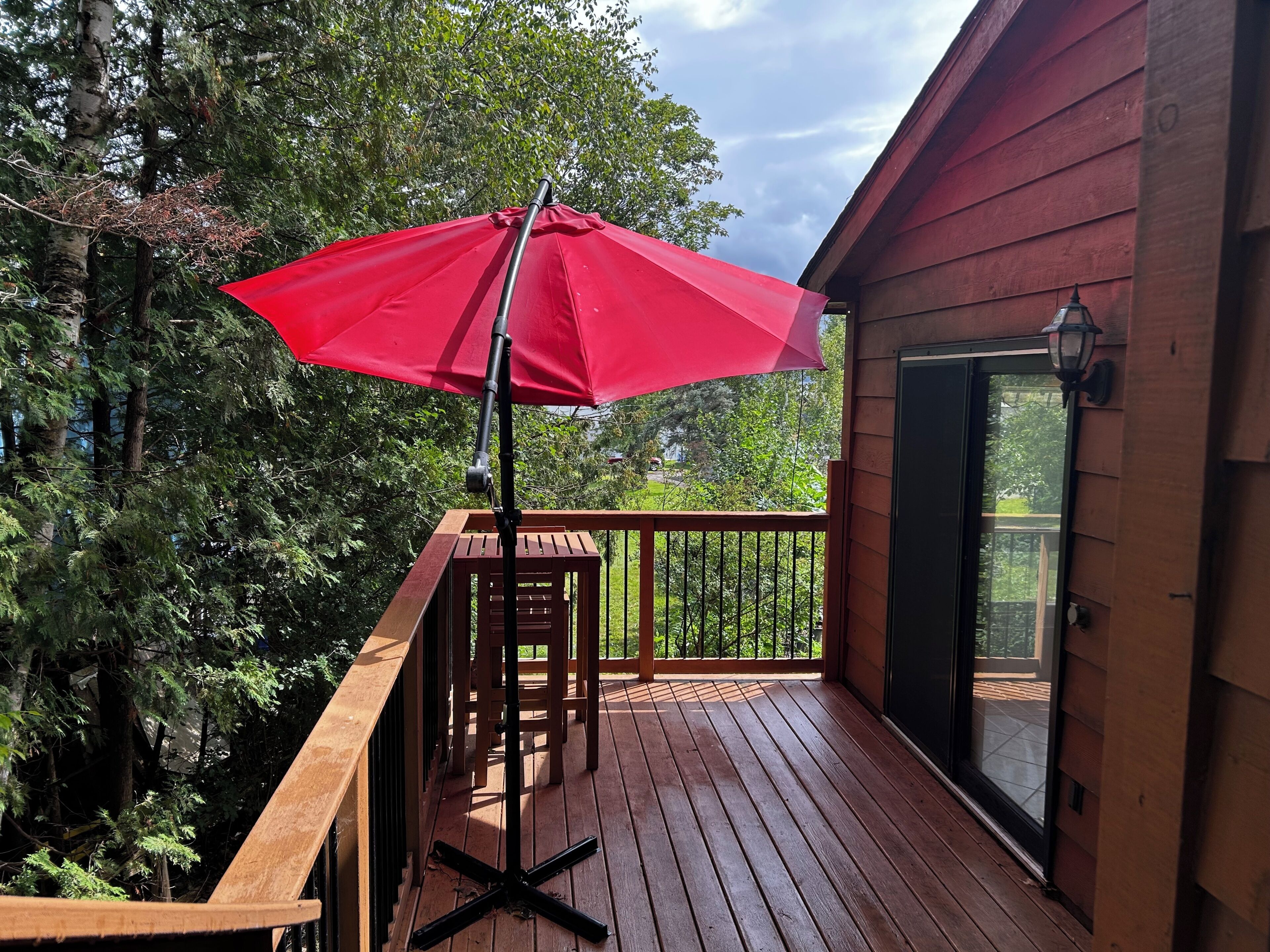 A wooden deck with a red umbrella, surrounded by trees and a building with a glass door.