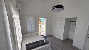 A bedroom with a bed, a bedside table, a hanging light fixture, a door, and a view of the outside.