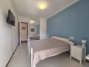 A bedroom with a bed, bedside table, lamp, and a TV mounted on the wall. - Holiday apartment with air conditioning (Bibione)
