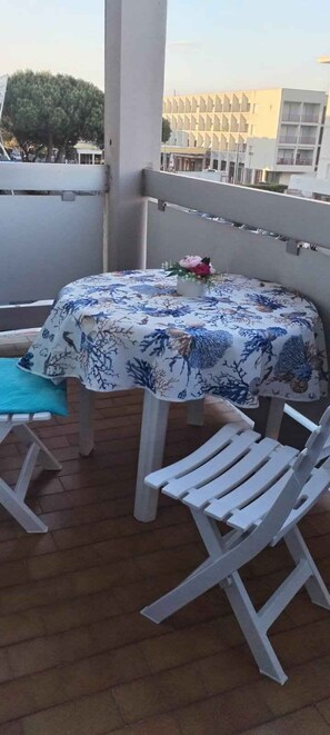 Un balcon avec une table recouverte d’une nappe à motifs floraux, deux chaises en plastique blanches et une petite plante en pot.