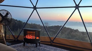 Una habitación con un ventanal grande, una estufa de leña, un ventilador y una alfombra estampada.