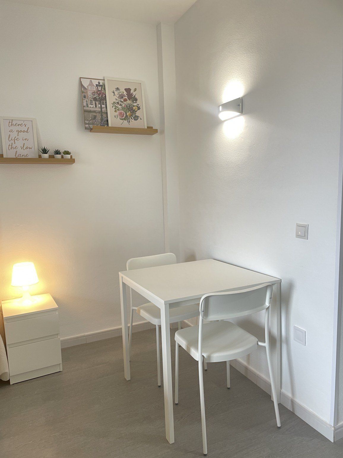 A room with a white table and chairs, a bedside table with a lamp, and wall-mounted shelves with framed pictures.