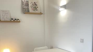 A room with a white table and chairs, a bedside table with a lamp, and wall-mounted shelves with framed pictures.