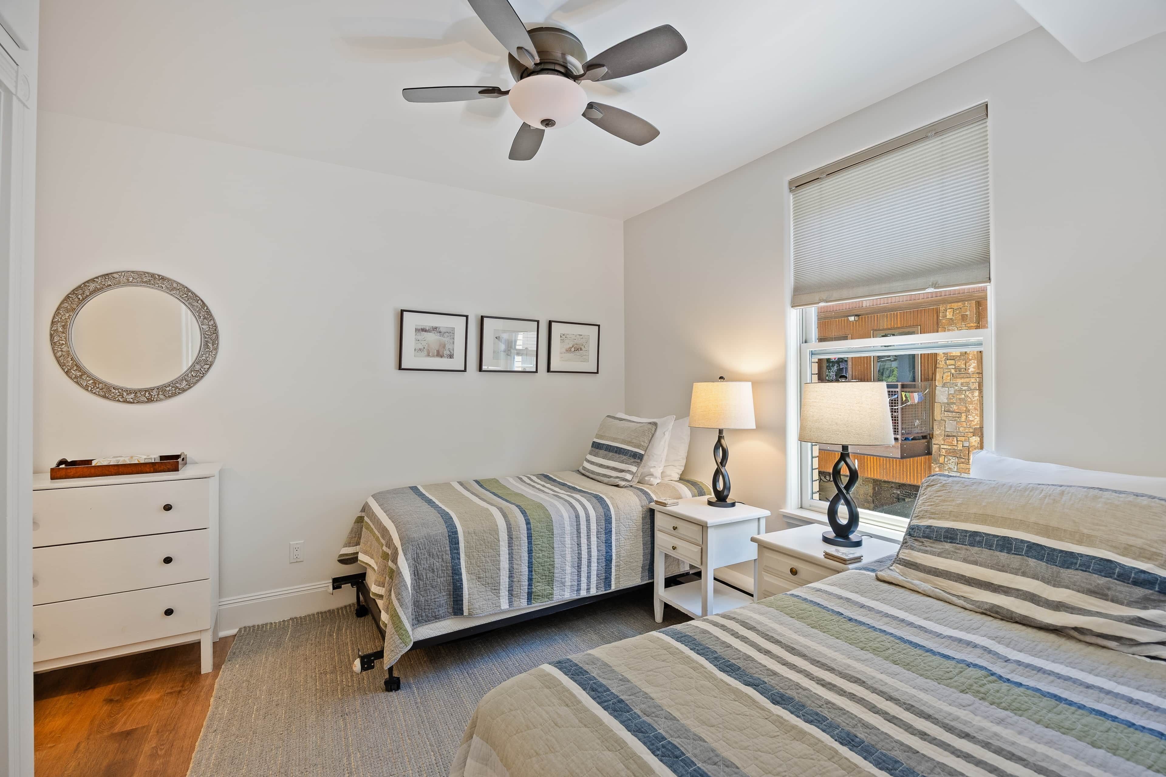 Une chambre à coucher comprenant un lit, une table de chevet, une lampe, un ventilateur de plafond, une commode et une fenêtre avec des stores.