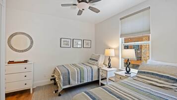 Une chambre à coucher comprenant un lit, une table de chevet, une lampe, un ventilateur de plafond, une commode et une fenêtre avec des stores.