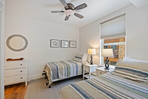 Ein Schlafzimmer mit einem Bett, einem Nachttisch, einer Lampe, einem Deckenventilator, einer Kommode und einem Fenster mit Jalousien.