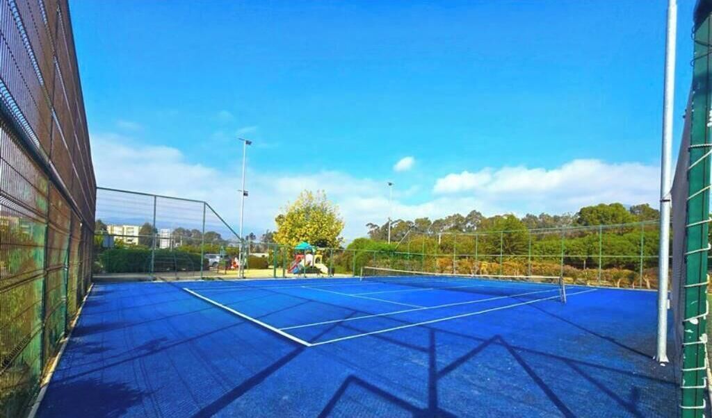 An indoor tennis court with a blue surface, surrounded by a metal fence, and trees in the background.