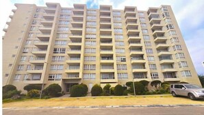 A multi-story residential building with balconies, a parking area, and a clear sky.
