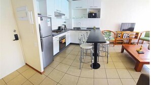A compact kitchen with white cabinets, a refrigerator, stove, and a microwave. There is a dining area with a table and chairs, and a television on a stand.