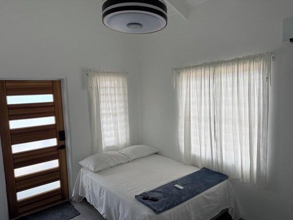 A single bed with a white mattress and a blue blanket, a wooden door with horizontal slats, and a ceiling light fixture.