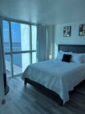 Ein Schlafzimmer mit großem Fenster, einem Bett mit weißer Bettwäsche, einem Nachttisch und zwei abstrakten Bildern an der Wand.