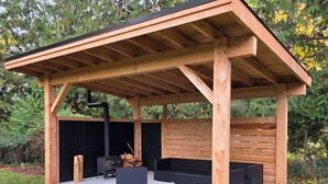 A covered outdoor seating area with a wooden structure, a black grill, and a firewood stack. - Halfmoon Bay Hideaway Top Suite (Halfmoon Bay)
