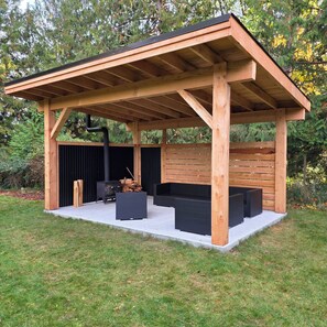Un espace extérieur abrité, doté d’une structure en bois, d’un barbecue noir et d’une pile de bois de chauffage.