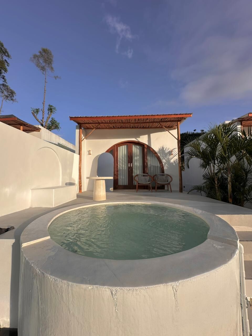 Un'area piscina con una vasca idromassaggio circolare, una zona relax coperta con sedie e un edificio con tetto in legno e portale ad arco.