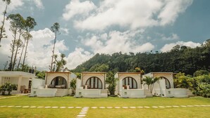 A row of white cottages with arched doorways, a grassy lawn, and a stone pathway leading to them. - Garden Hill Kintamani Natural Hot Spring (Kintamani)