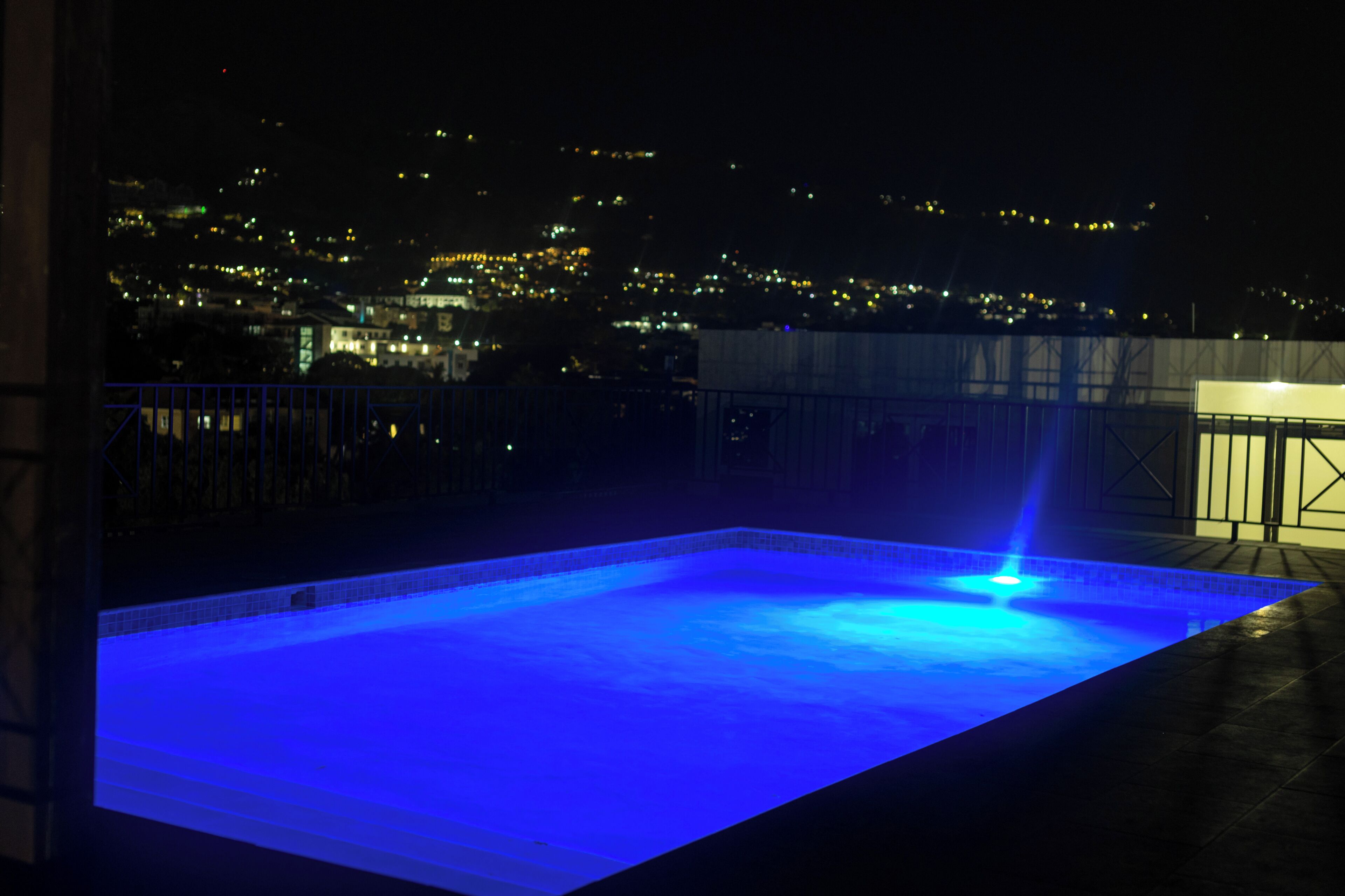 Une piscine rectangulaire dont l’eau est illuminée de bleu, agrémentée d’un élément décoratif aquatique, et entourée d’une rambarde métallique, offrant une vue sur le paysage urbain la nuit.