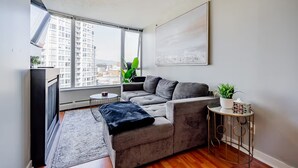 A modern living room with a grey sofa, a small side table, a large window with a view of buildings, and a ceiling light. - 1BR Condo Near Rogers Arena | Heart of Vancouver (Vancouver)